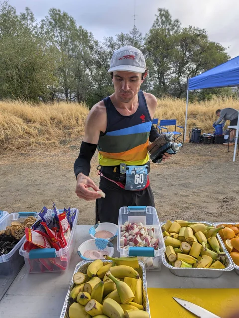 Runner in a white cap and striped singlet choosing food from an aid-station table with bananas and snacks.