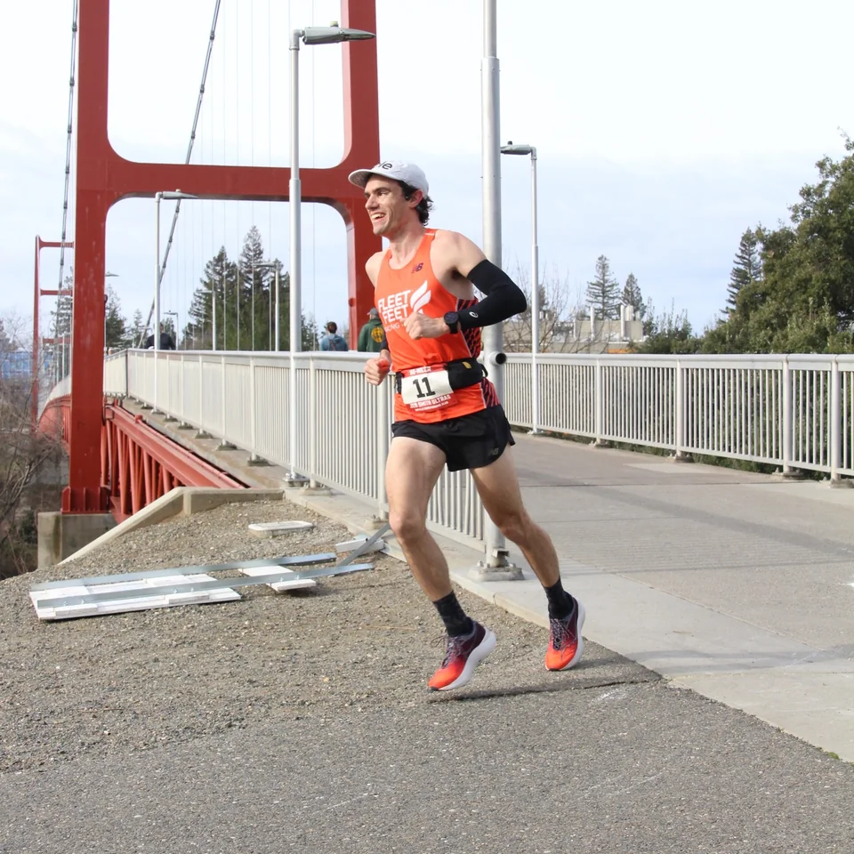 Brandon Costa running beside a red suspension bridge in an orange singlet.