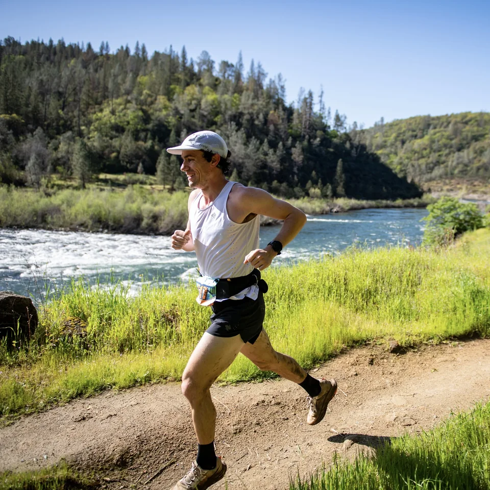 Brandon Costa running beside a river in a white singlet and cap.