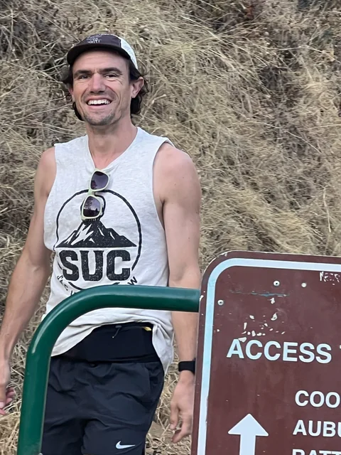 Brandon Costa smiling in a cap and gray SUC shirt beside a trail sign and green railing.