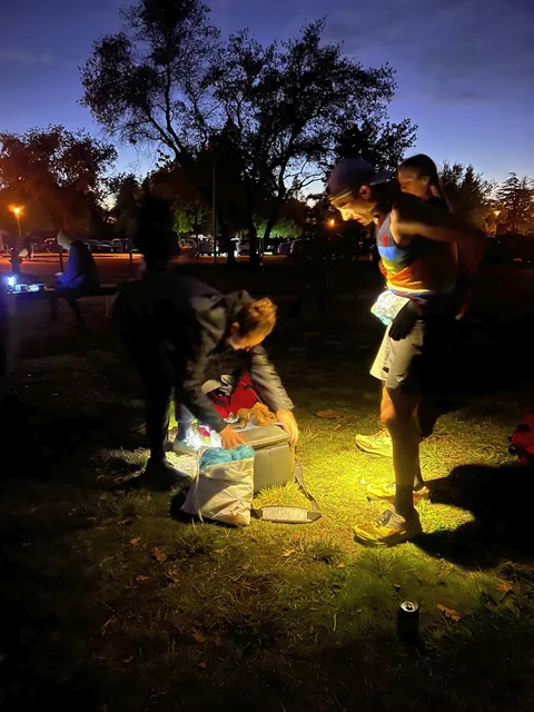 Runner in a striped singlet standing beside another person crouching over supplies outdoors at dusk.