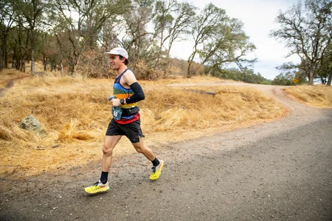 Runner in a white cap and striped singlet striding along a road beside dry grass and oak trees.