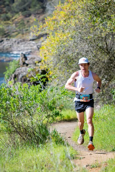 Runner in a white singlet approaching on a narrow path bordered by green plants beside the river.