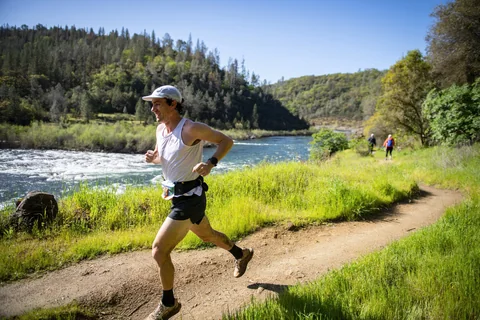 Runner in a white singlet and cap on a dirt trail beside a river and wooded hills.