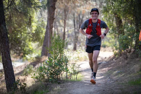 Runner wearing a red hydration vest on a shaded dirt trail through woodland.