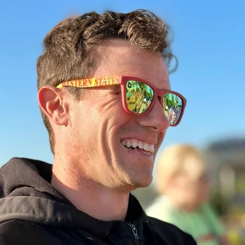 Close-up of a smiling man wearing mirrored sunglasses beneath a blue sky.