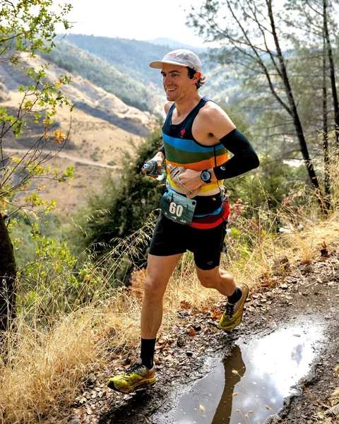 Brandon Costa smiling as he runs beside a puddle on a trail above a wooded valley.