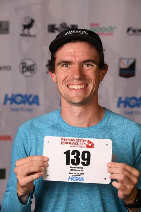 Brandon Costa smiling and holding a Western States race bib.